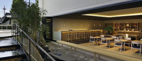 An outdoor seating area with tables and chairs, a small garden with bamboo, and a view of a traditional building in the background.
