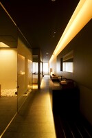 A modern hotel corridor with a glass partition, a reception desk, and a bed in the distance.
