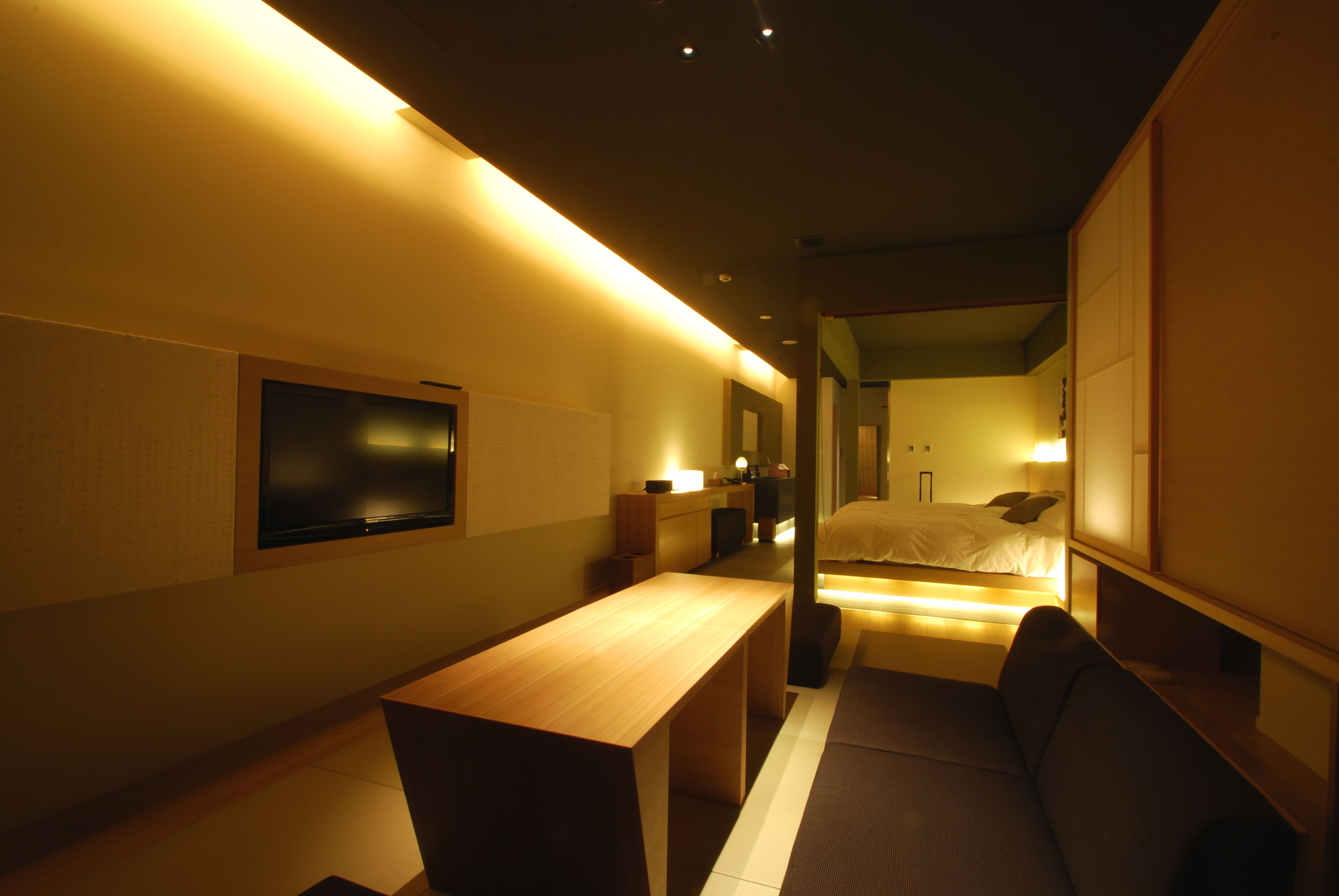 A modern hotel room with a wooden desk, a sofa, a bed, and a TV mounted on the wall.