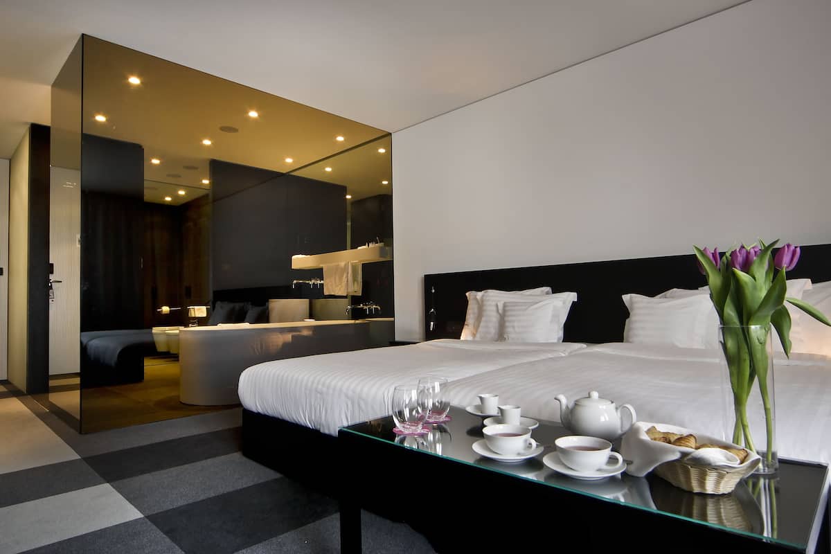 A modern hotel room with a large bed, a glass table with breakfast items, a vase of flowers, and a mirror reflecting the room.
