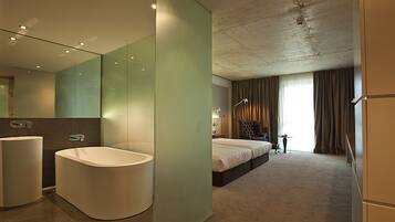 A modern hotel room with a large bathtub, a separate bedroom with a bed, a chair, and a desk.