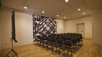 Une salle de conférence avec des rangées de chaises, un support pour projecteur et un motif géométrique sur le mur.