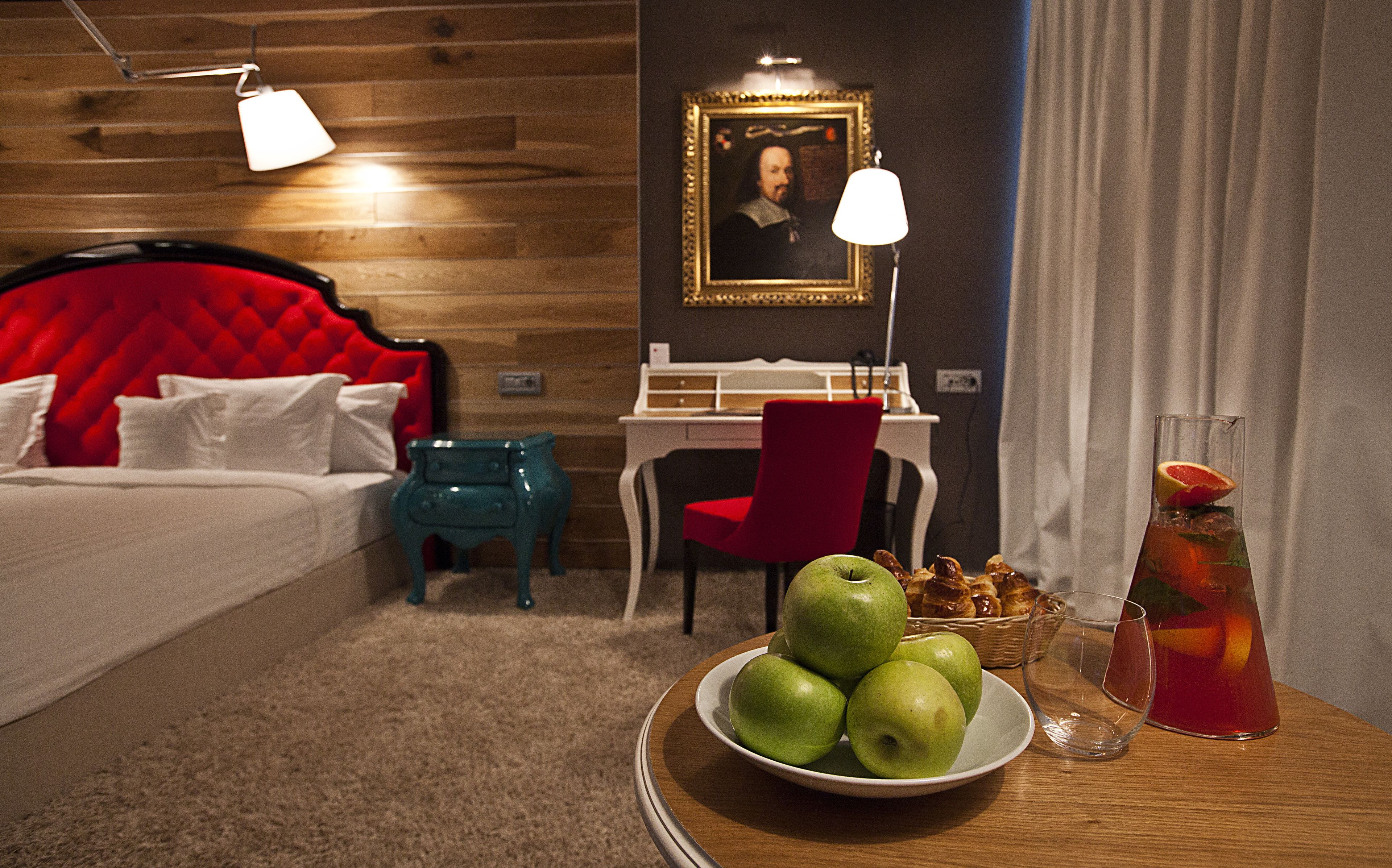 A hotel room with a red upholstered headboard, a wooden desk, a red chair, a white lamp, a framed portrait on the wall, a bowl of green apples, and a glass of red drink on a wooden table.