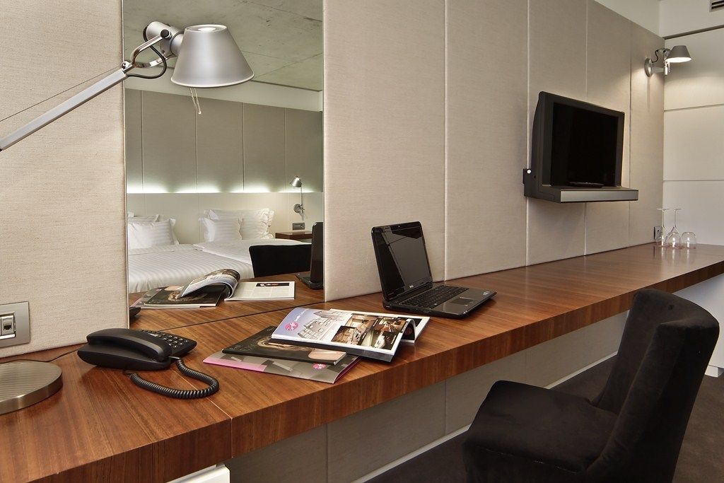Habitación de hotel con un escritorio de madera, una computadora portátil, un teléfono y una revista. Hay un televisor montado en la pared y una cama con sábanas blancas al fondo.
