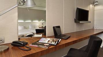 Habitación de hotel con un escritorio de madera, una computadora portátil, un teléfono y una revista. Hay un televisor montado en la pared y una cama con sábanas blancas al fondo.