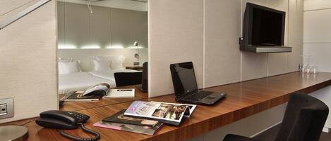 A hotel room with a wooden desk, a laptop, a phone, and a magazine. There is a TV mounted on the wall and a bed with white linens in the background.