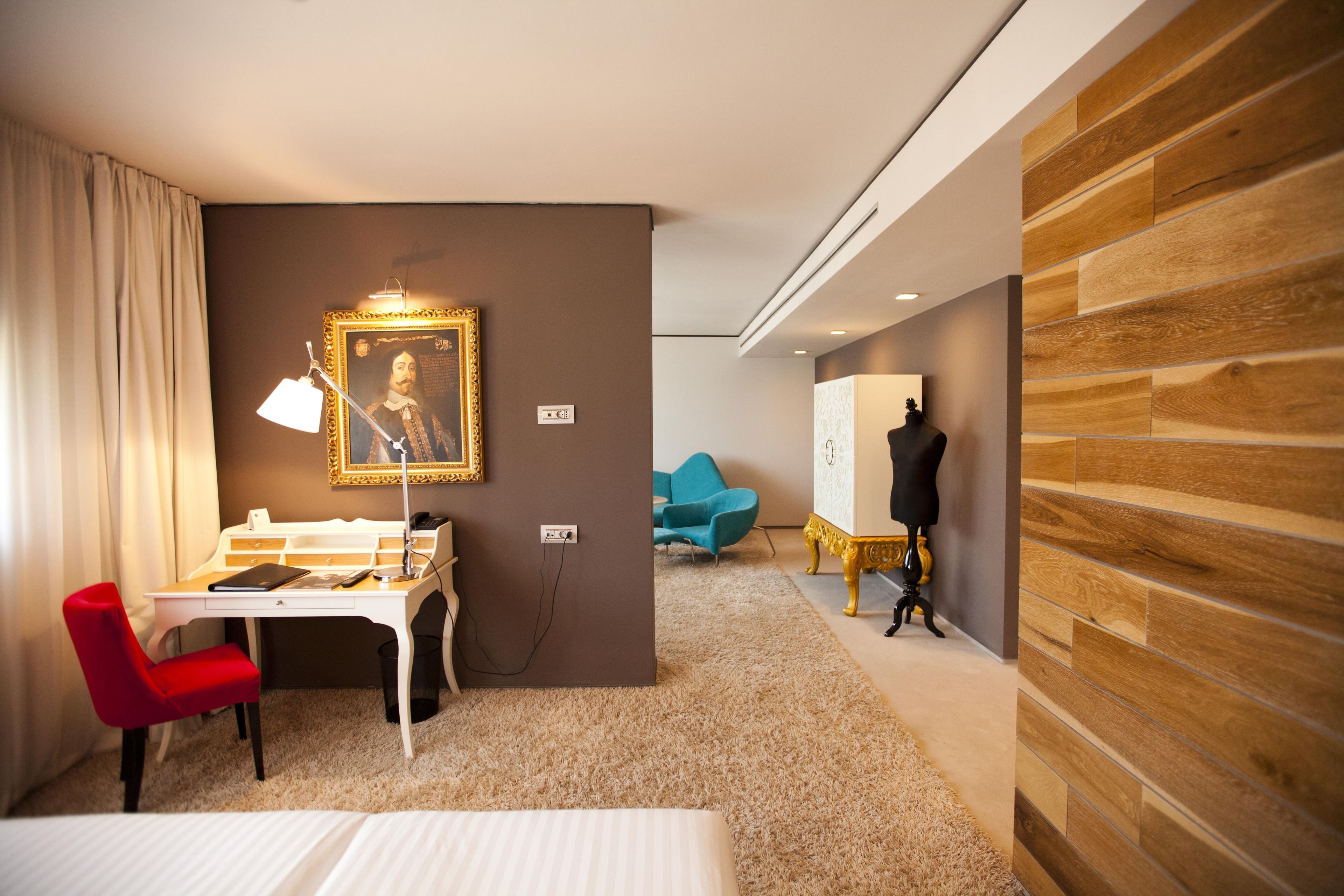 Habitación de hotel con escritorio, una silla roja, cama, un retrato en la pared y un maniquí vestido de negro.