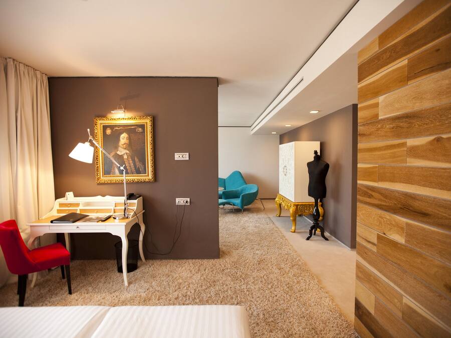 Habitación de hotel con escritorio, una silla roja, cama, un retrato en la pared y un maniquí vestido de negro.