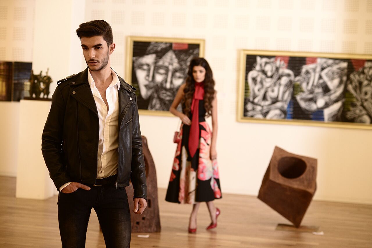Un hombre con chaqueta de cuero y una mujer con vestido floral en una galería de arte con pinturas abstractas.