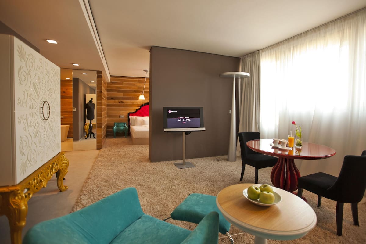 A modern hotel room with a TV, a dining table with chairs, a sofa, and a staircase leading to another room.