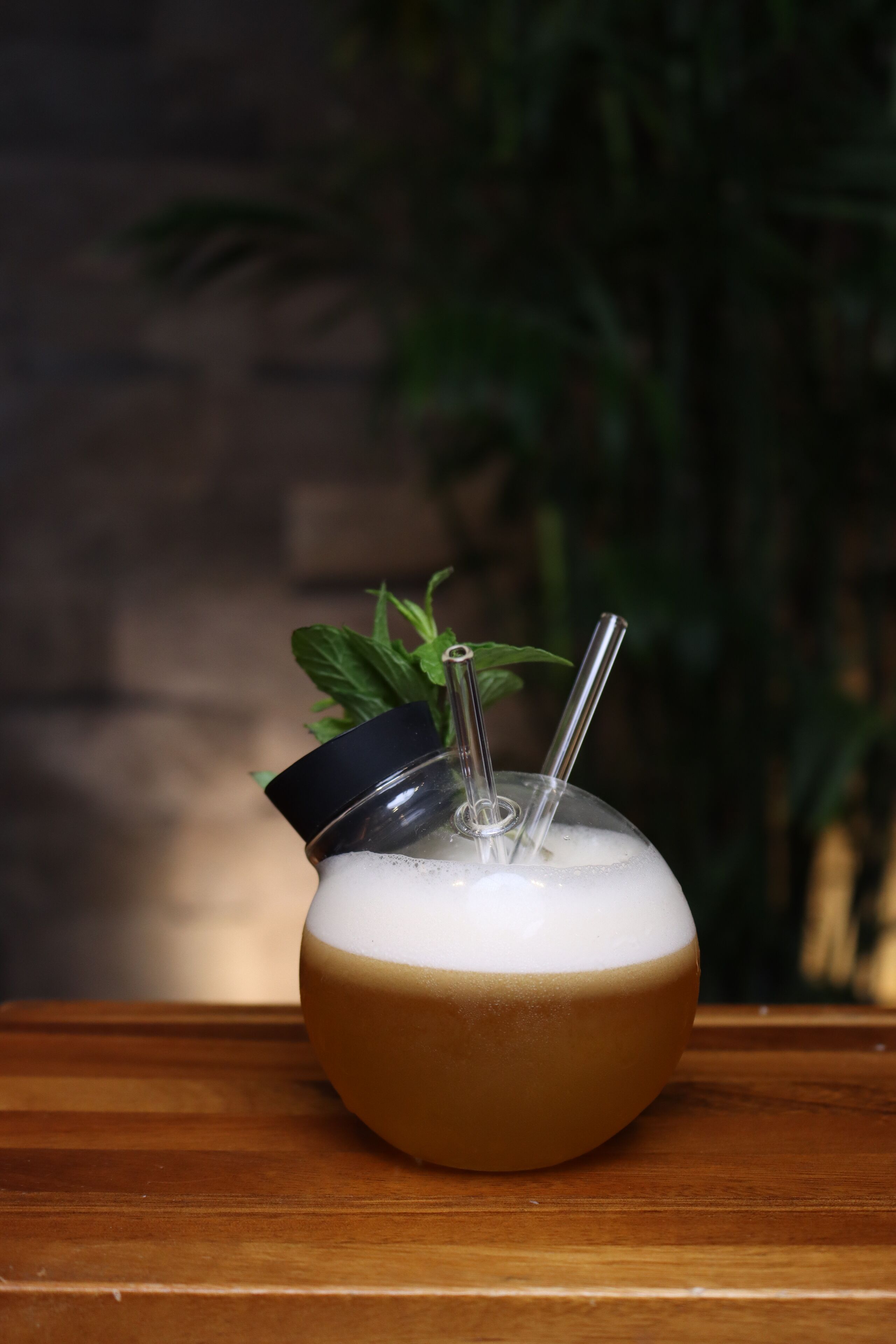 Una taza de vidrio transparente con tapa negra y una pajita, llena de una bebida espumosa, sobre una superficie de madera con una planta verde desenfocada al fondo.