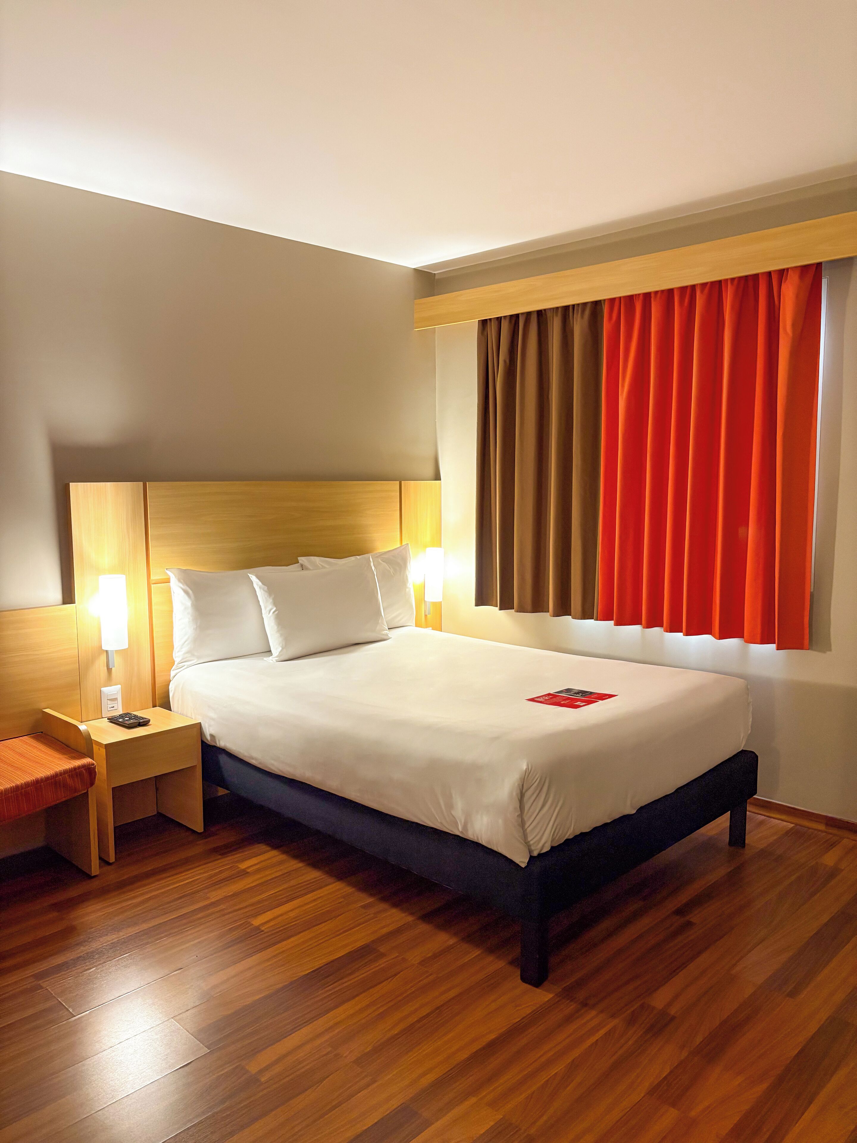 A hotel room with a wooden headboard, a bed with white linens, a nightstand with a lamp, and a window with orange curtains.
