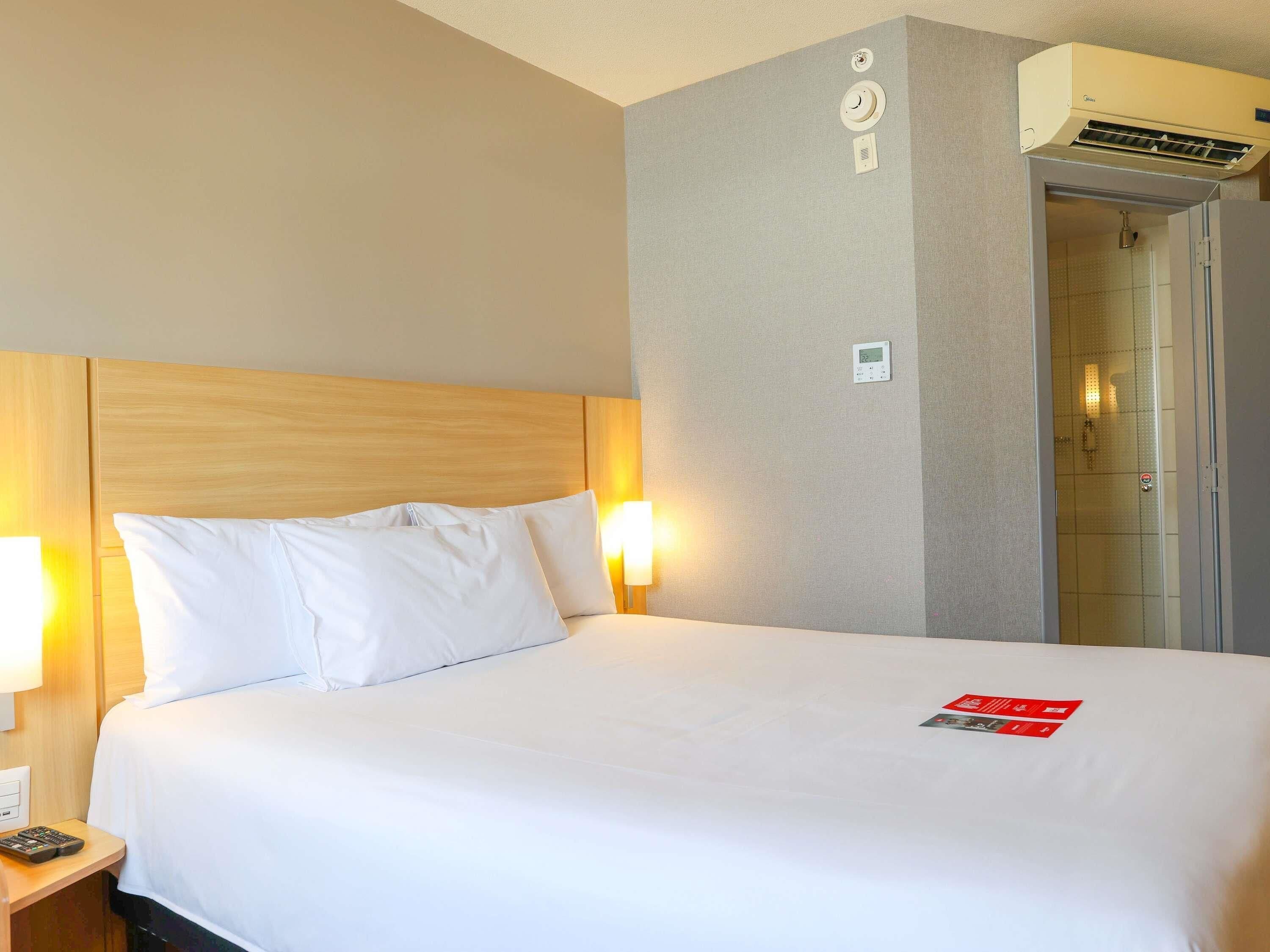 Une chambre d’hôtel équipée d’un lit, de lampes de chevet, d’un appareil de climatisation et d’un panneau de commande mural.