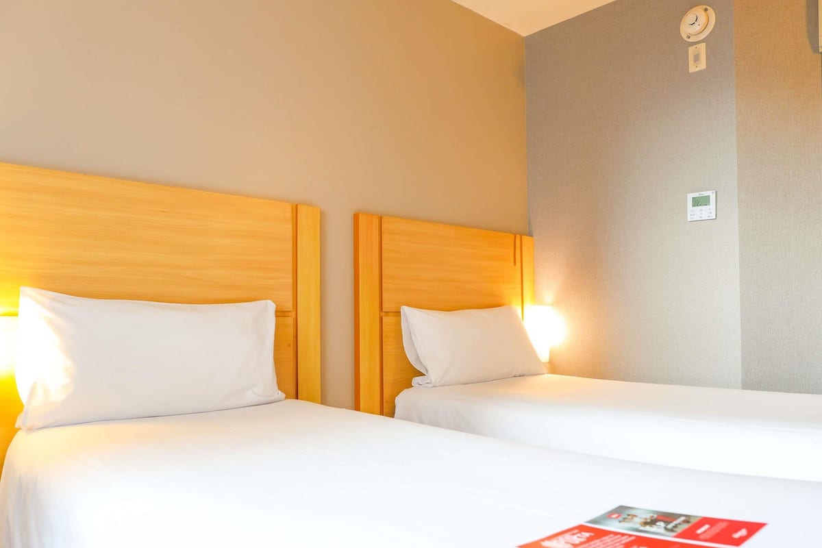 A hotel room with two beds, each with a wooden headboard and white bedding. There is a wall-mounted thermostat and a small clock on the wall.