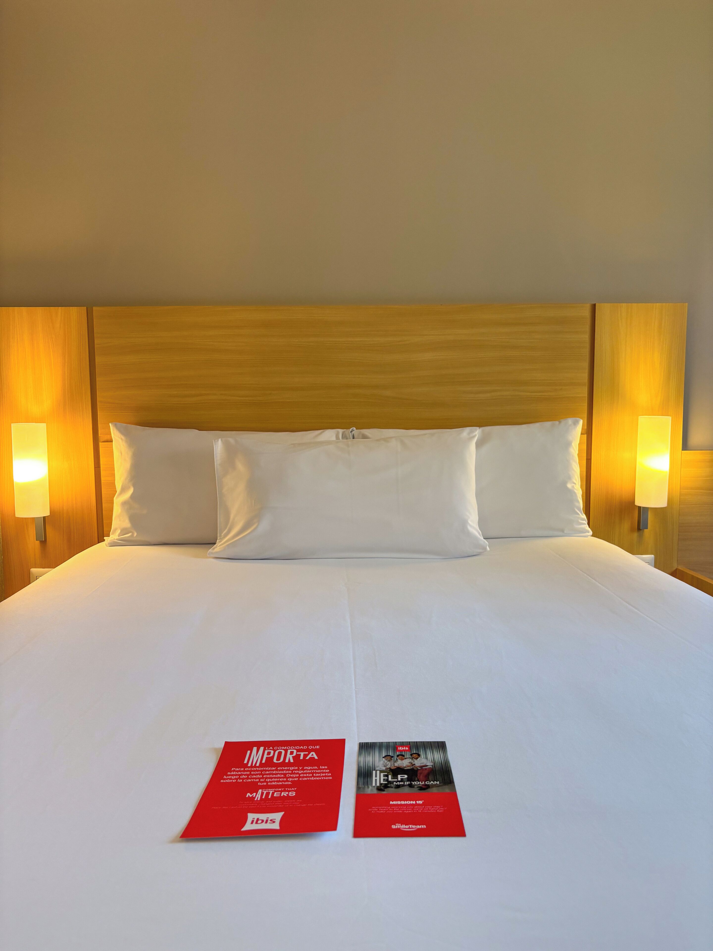 A hotel room with a large bed, wooden headboard, two bedside lamps, and two informational pamphlets on the bed.