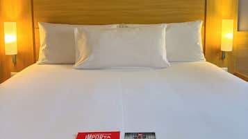 Habitación de hotel con una cama grande, cabecera de madera, dos lámparas de noche y dos folletos informativos sobre la cama.