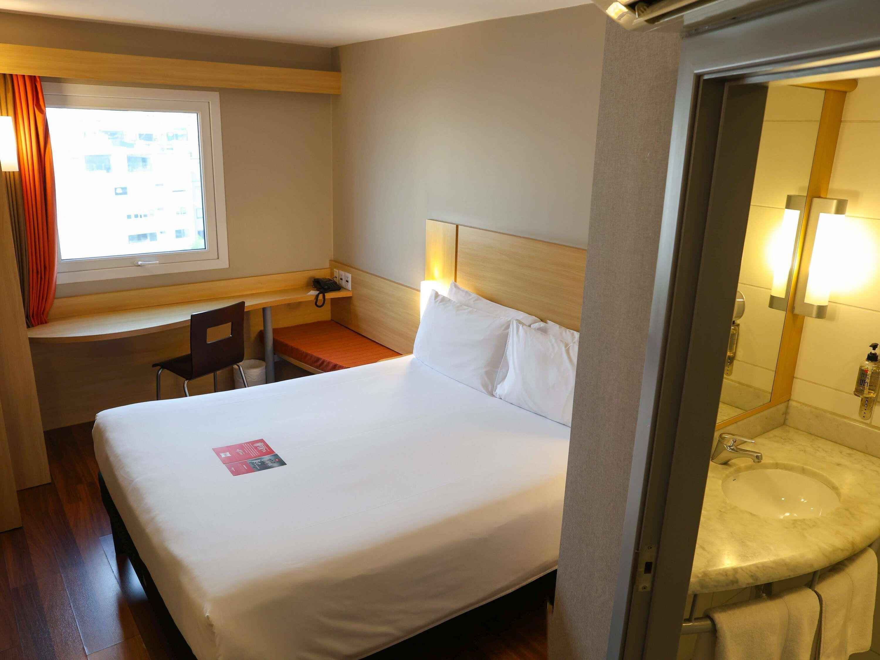 Habitación de hotel con cama, escritorio, silla, ventana con cortinas y baño con lavamanos y espejo.