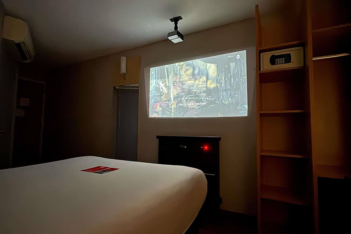 A hotel room with a bed, a projector screen, a desk, and a shelf.
