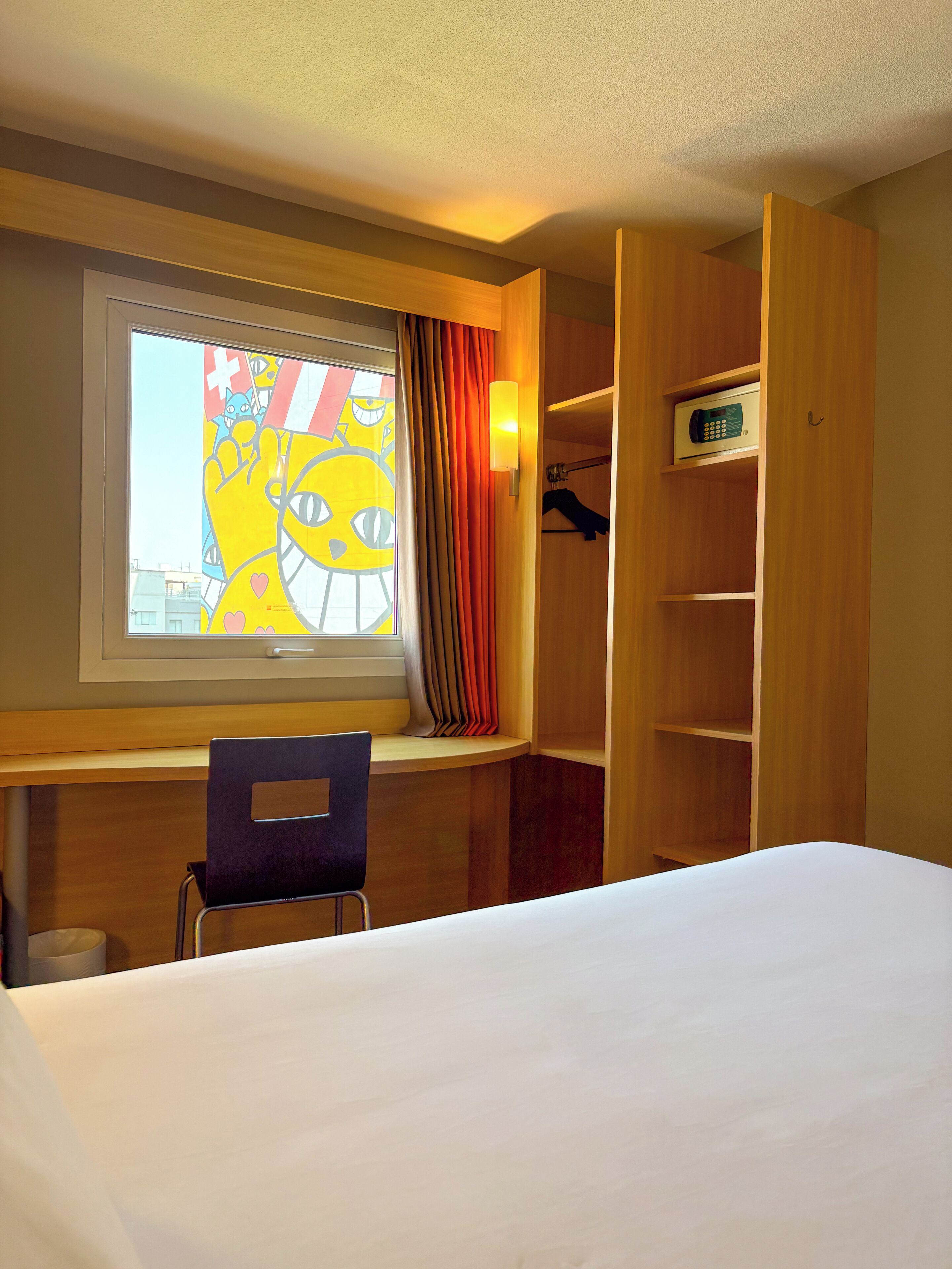 A hotel room with a bed, a chair, a window with a view, a wardrobe, and a wall-mounted light.