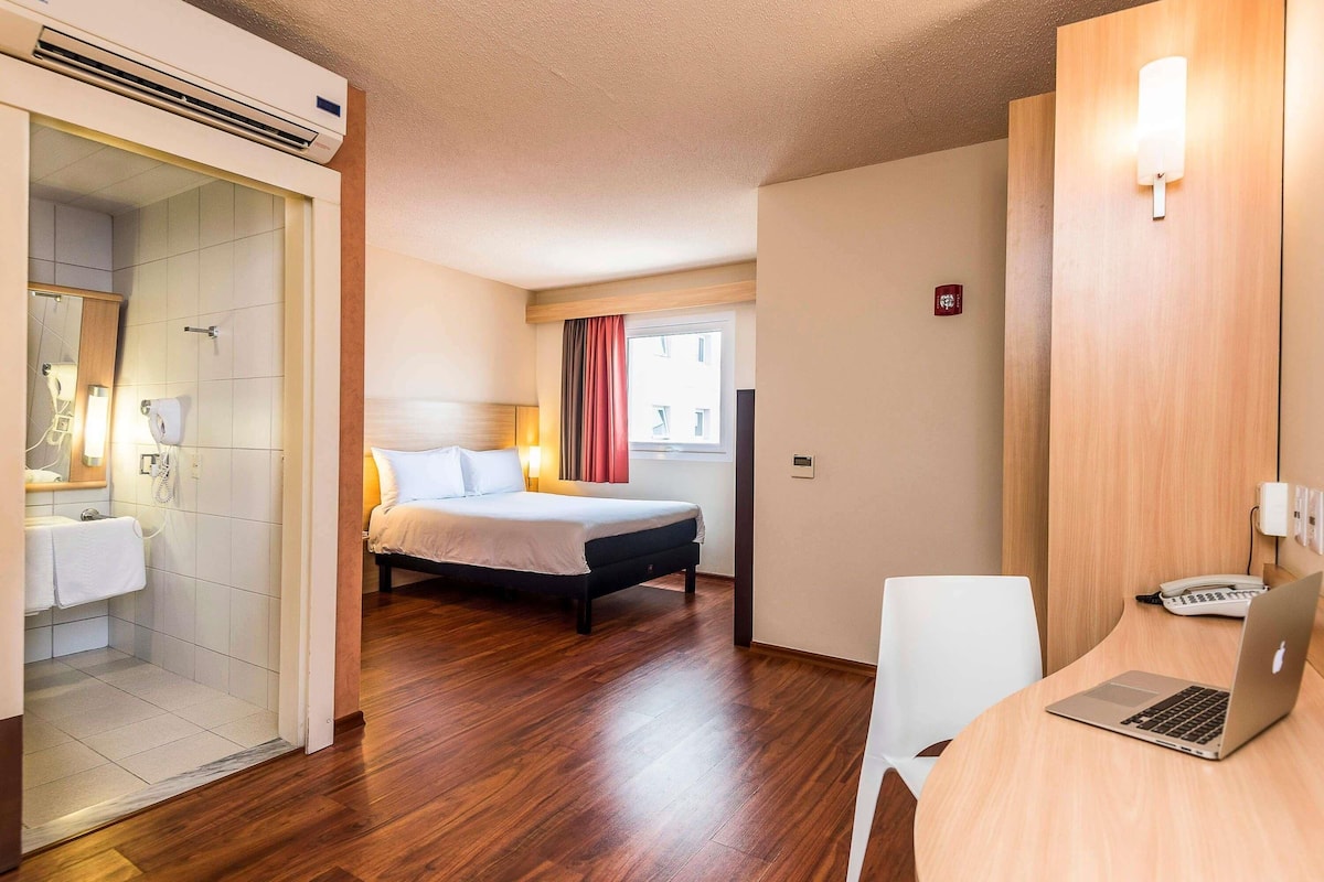 A hotel room with a bed, a desk with a laptop, a shower area, and a bathroom with a mirror and towel rack.