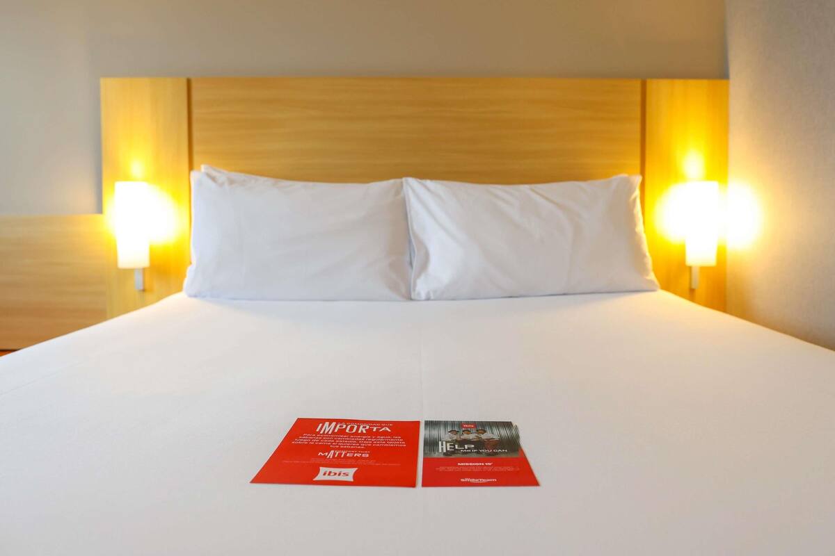 A hotel room with a wooden headboard, two bedside lamps, and a bed with white linens. There are two informational cards on the bed.