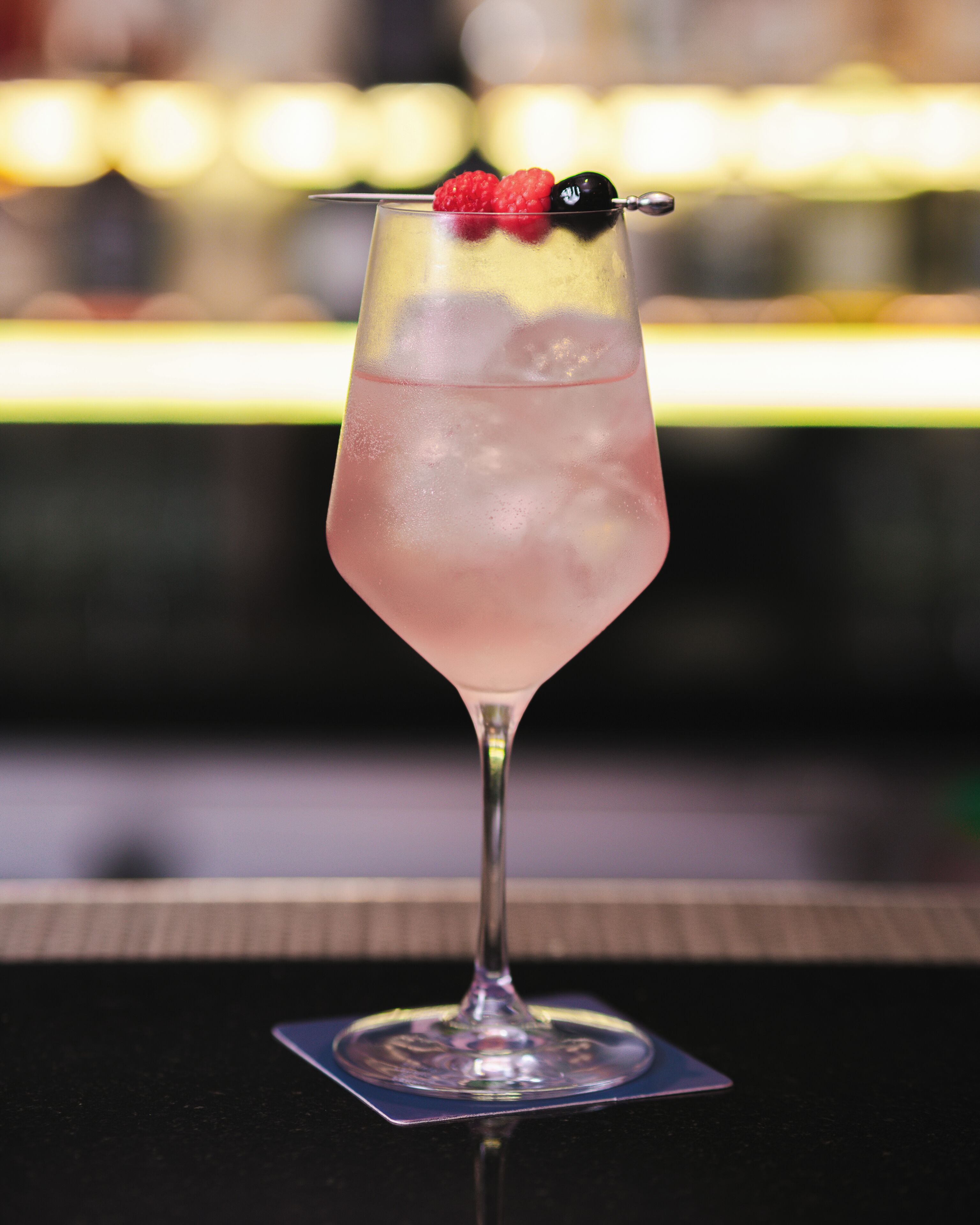 A cocktail with a raspberry garnish in a tall glass on a bar counter.