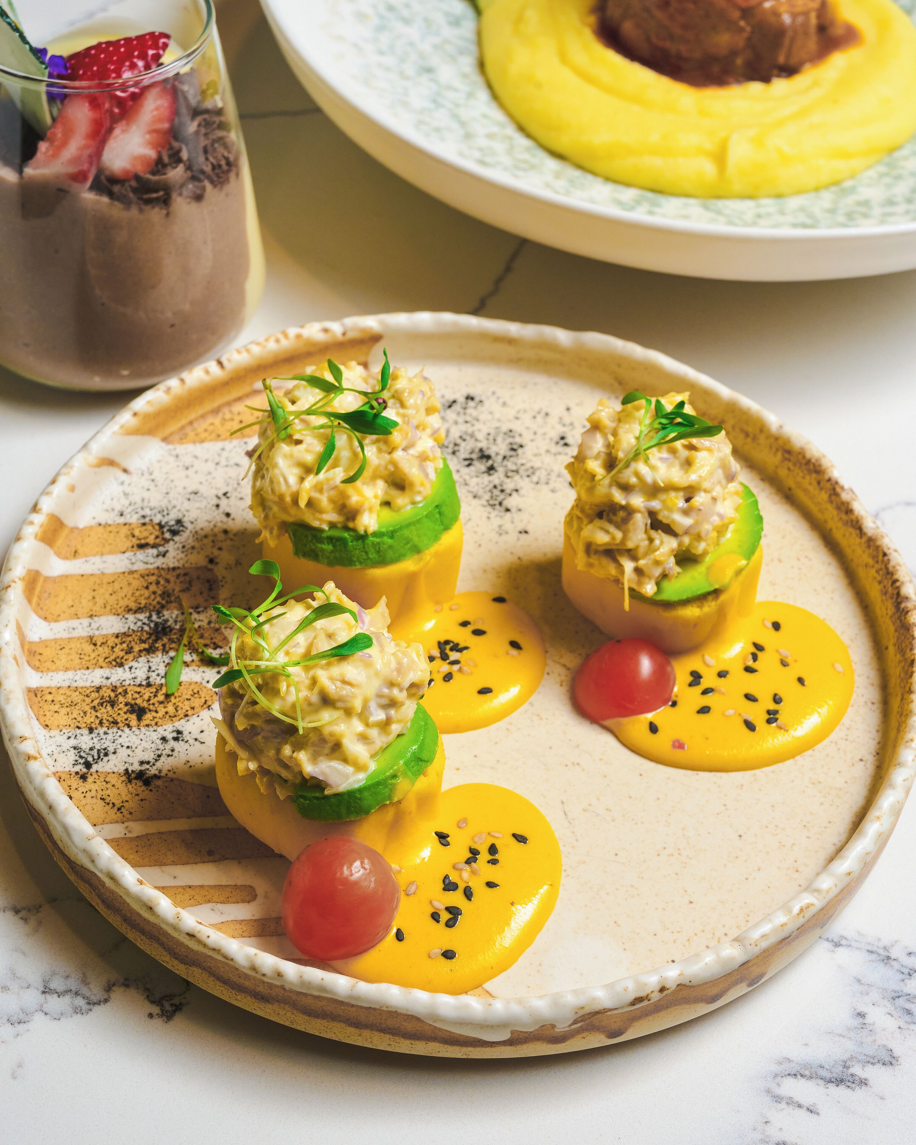 A plate of stuffed avocados with a creamy filling, garnished with herbs and black sesame seeds, accompanied by a small cherry tomato and a slice of mango.