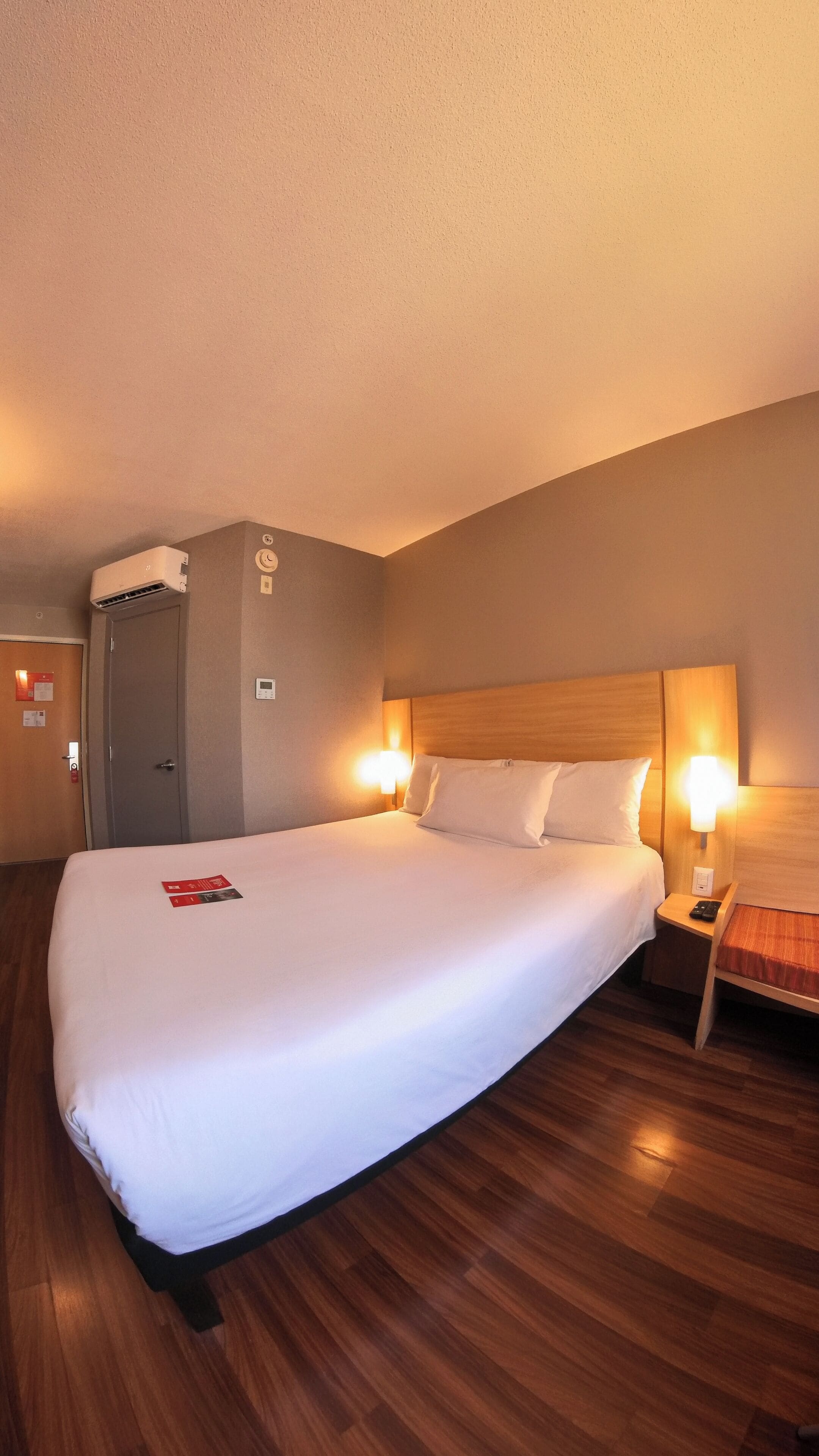 Une chambre d’hôtel avec un grand lit, une tête de lit en bois, des lampes de chevet et un climatiseur fixé au mur.