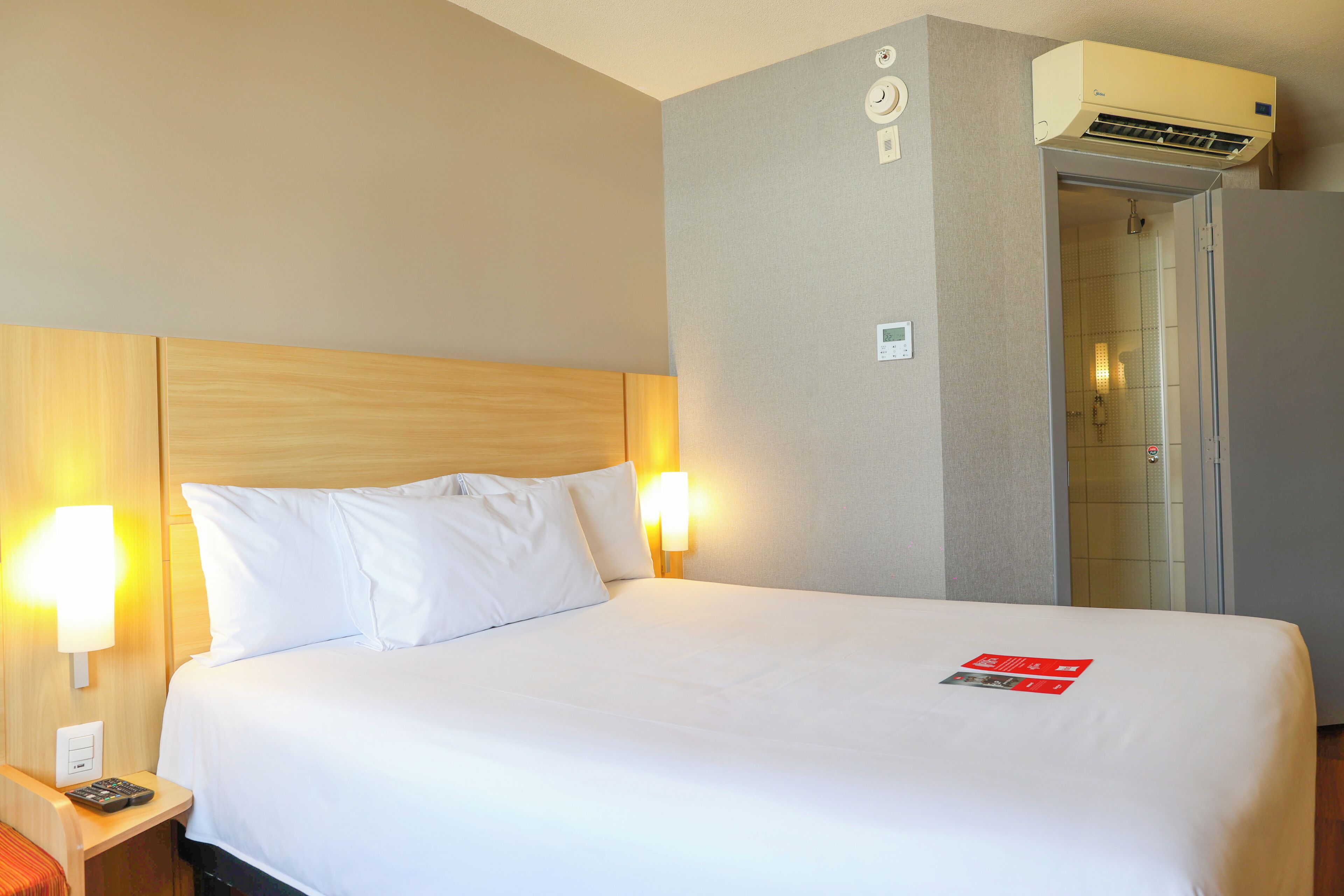 Een hotelkamer met een bed, nachtlampjes, een bedieningspaneel en een airconditioning.
