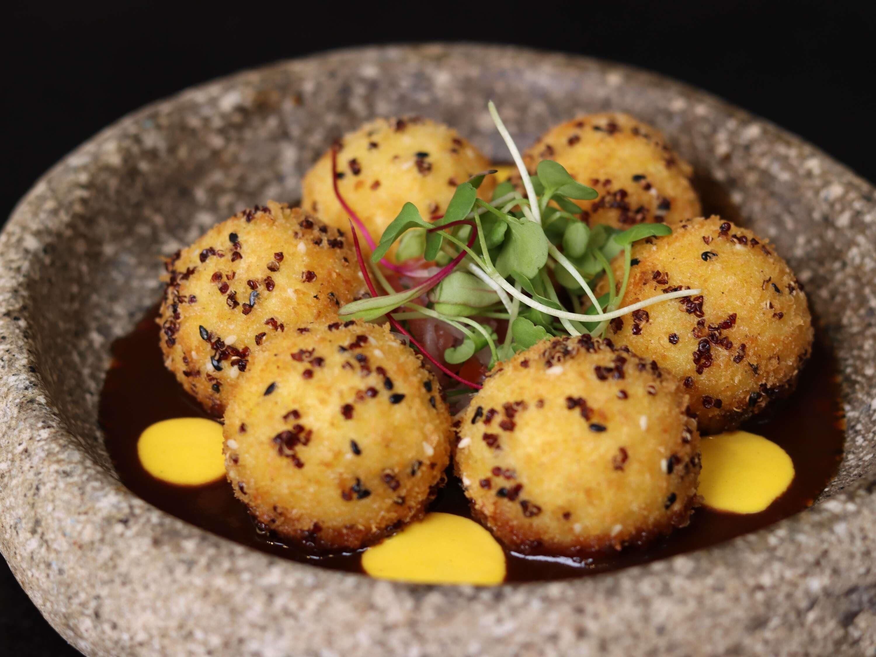 A dish of round, breaded balls garnished with microgreens and served in a stone bowl with a dark sauce.