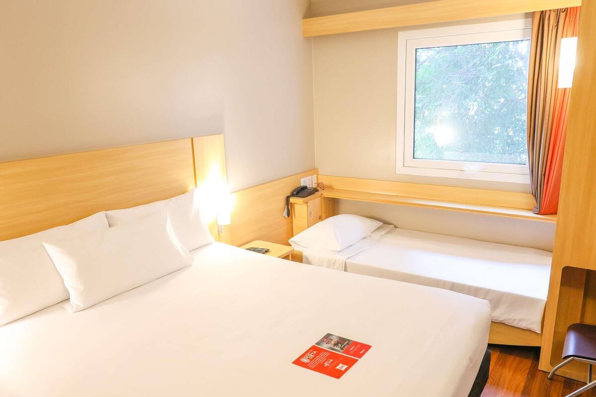 A hotel room with a large bed, a smaller bed, a bedside table with a phone, a window with curtains, and a wooden headboard.
