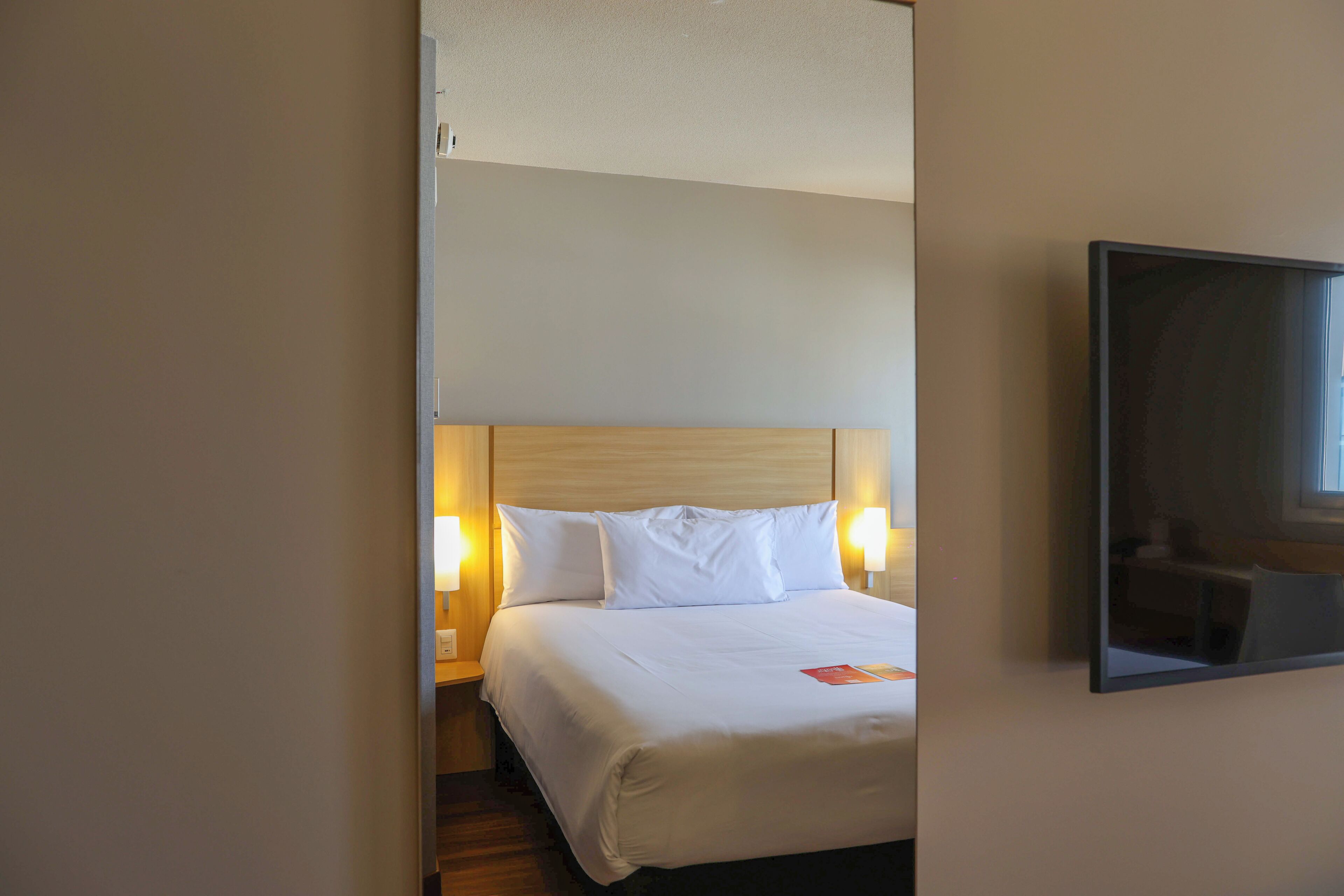 Ein Hotelzimmer mit Bett, Nachttischlampen, einem Nachttisch und einem an der Wand befestigten Fernseher.