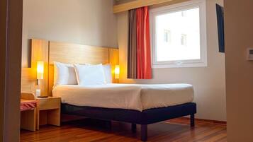 Habitación de hotel con suelo de madera, cama con ropa de cama blanca, mesita de noche con lámpara y ventana con cortinas.