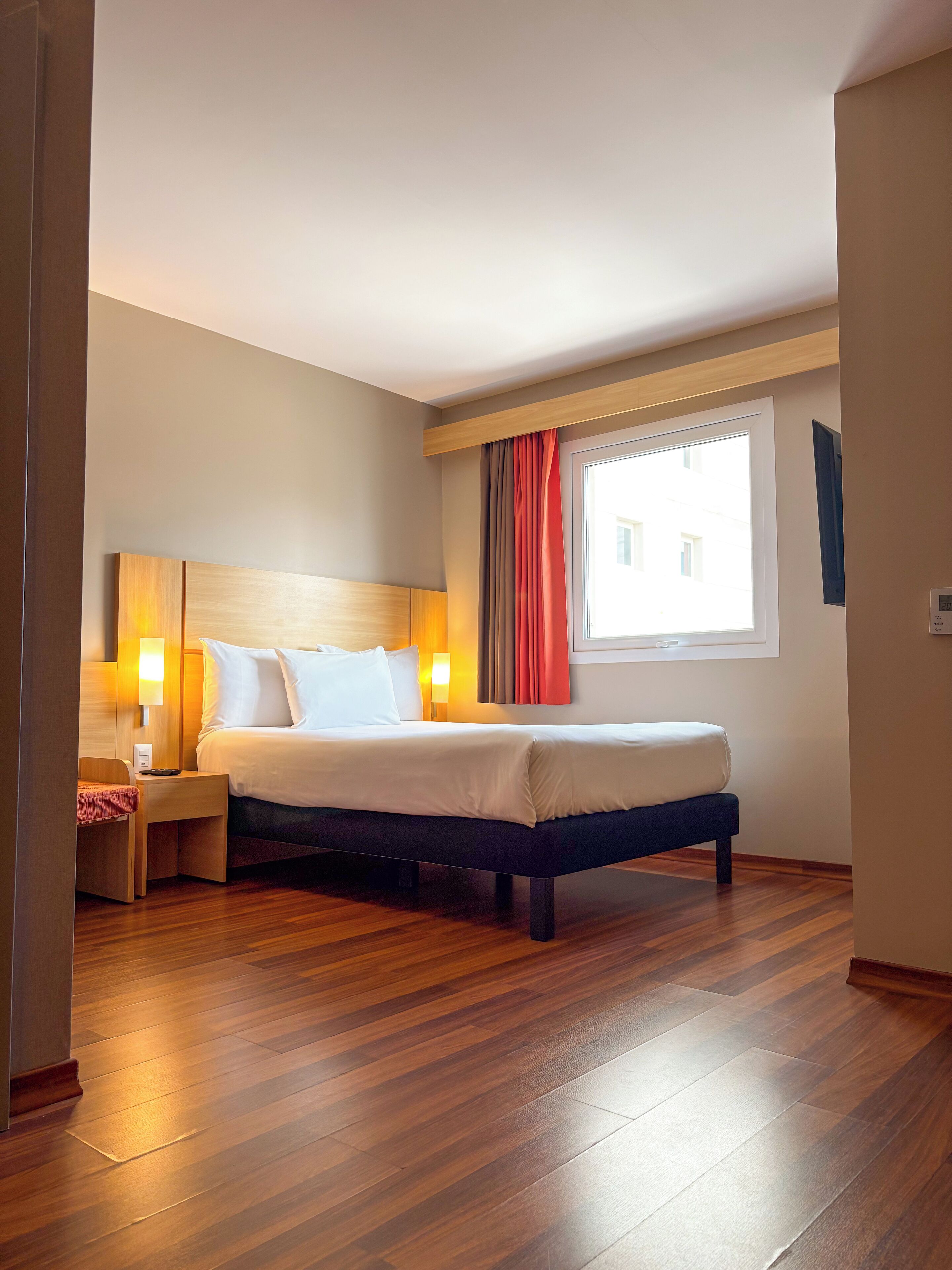 A hotel room with a wooden floor, a bed with white linens, a nightstand with a lamp, and a window with curtains.