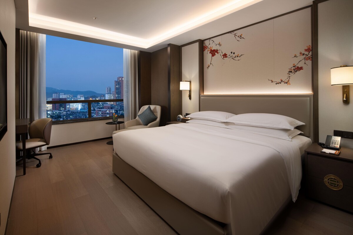 Habitación de hotel con una cama grande, un escritorio, una silla, una mesita de noche, una lámpara y vistas a la ciudad.