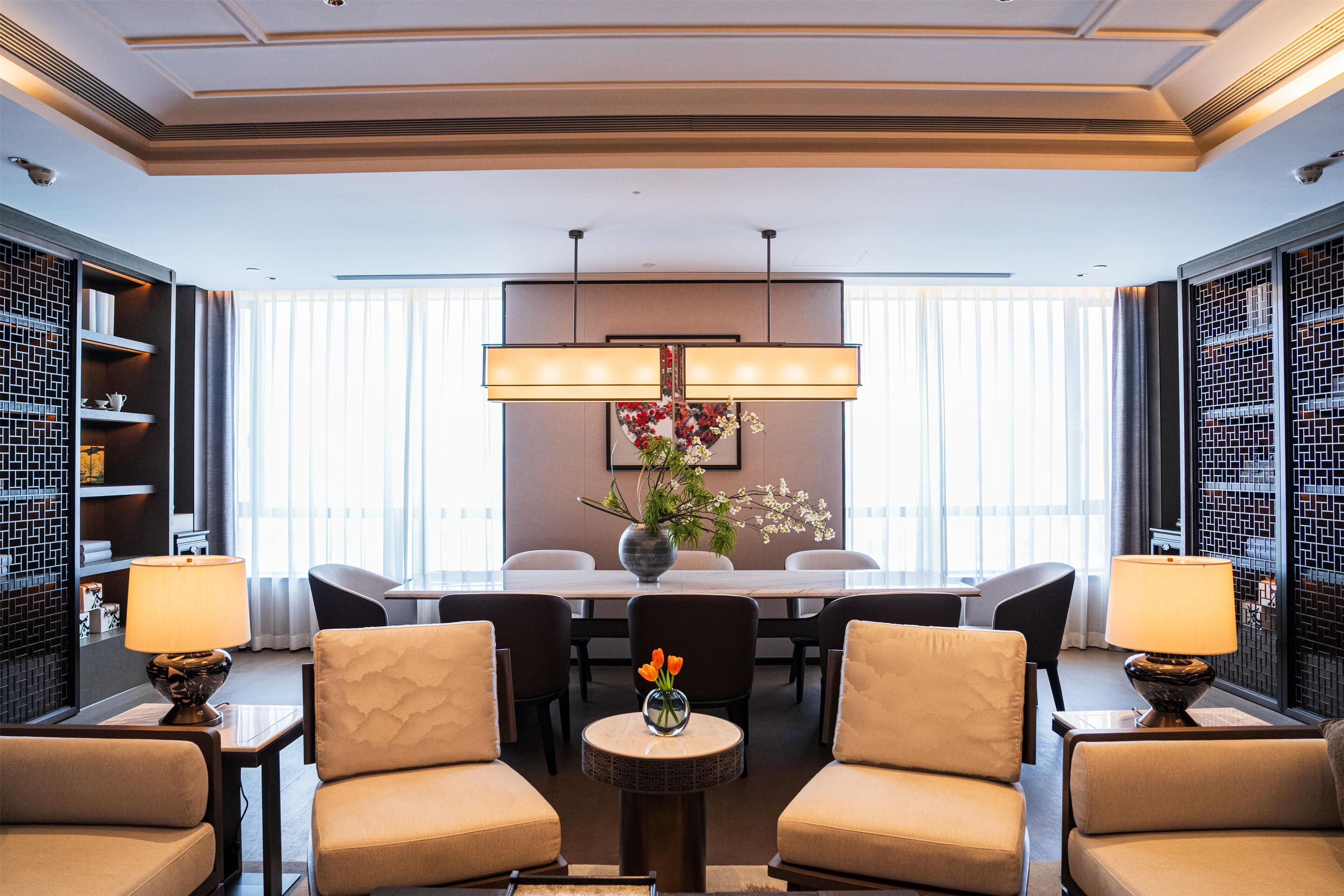 A hotel lobby with a dining area, two sofas, a round table with a vase of flowers, and a large chandelier.