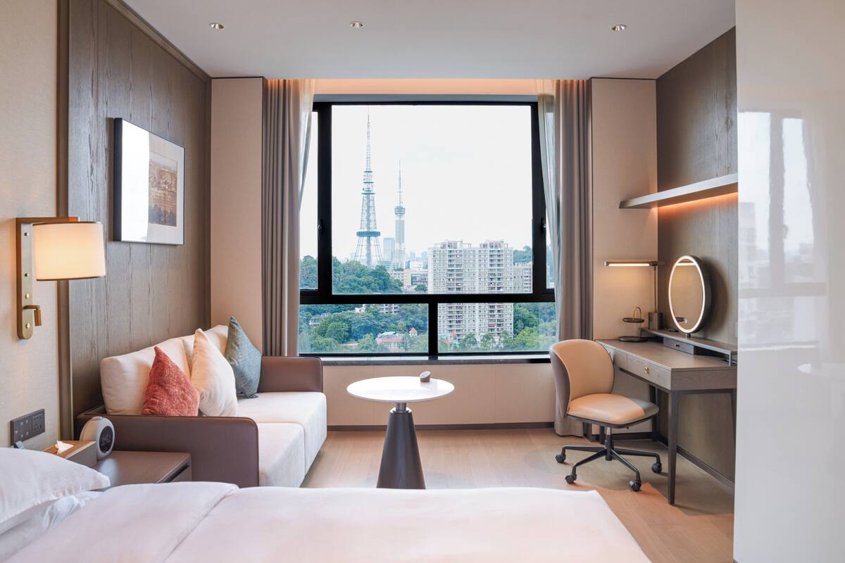 A hotel room with a bed, a desk, a chair, a small table, and a view of a cityscape through a large window.