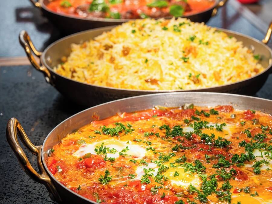 A variety of dishes in pans, including a pasta dish, a rice dish, and a tomato-based dish with herbs.