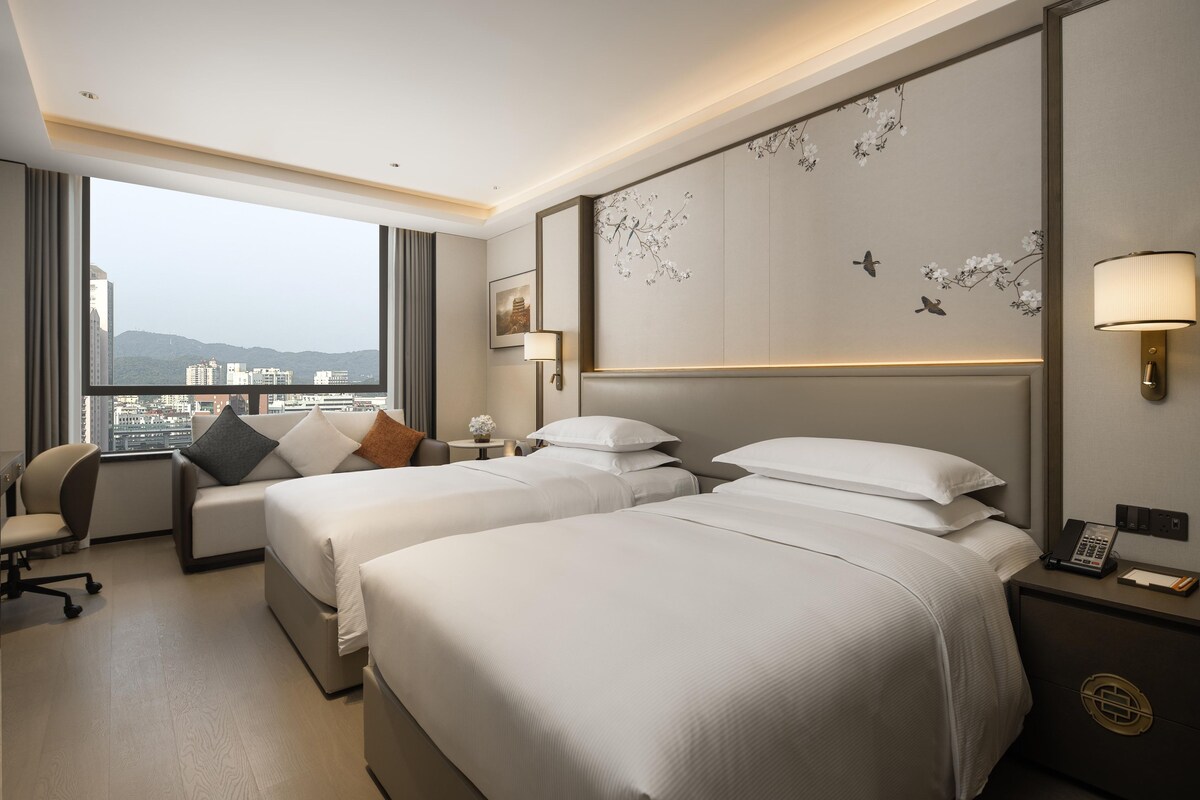 Habitación de hotel con dos camas, un escritorio y vistas a la ciudad.