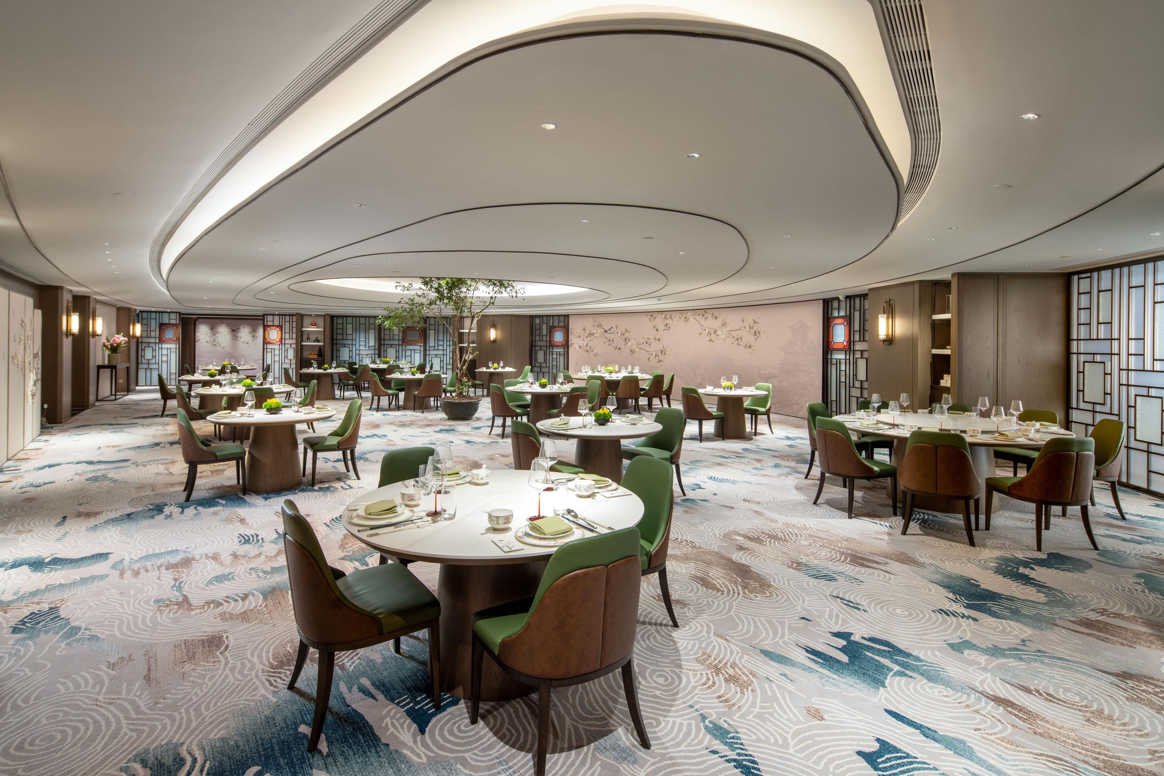 A spacious hotel dining area with multiple round tables, green upholstered chairs, and a patterned carpet.