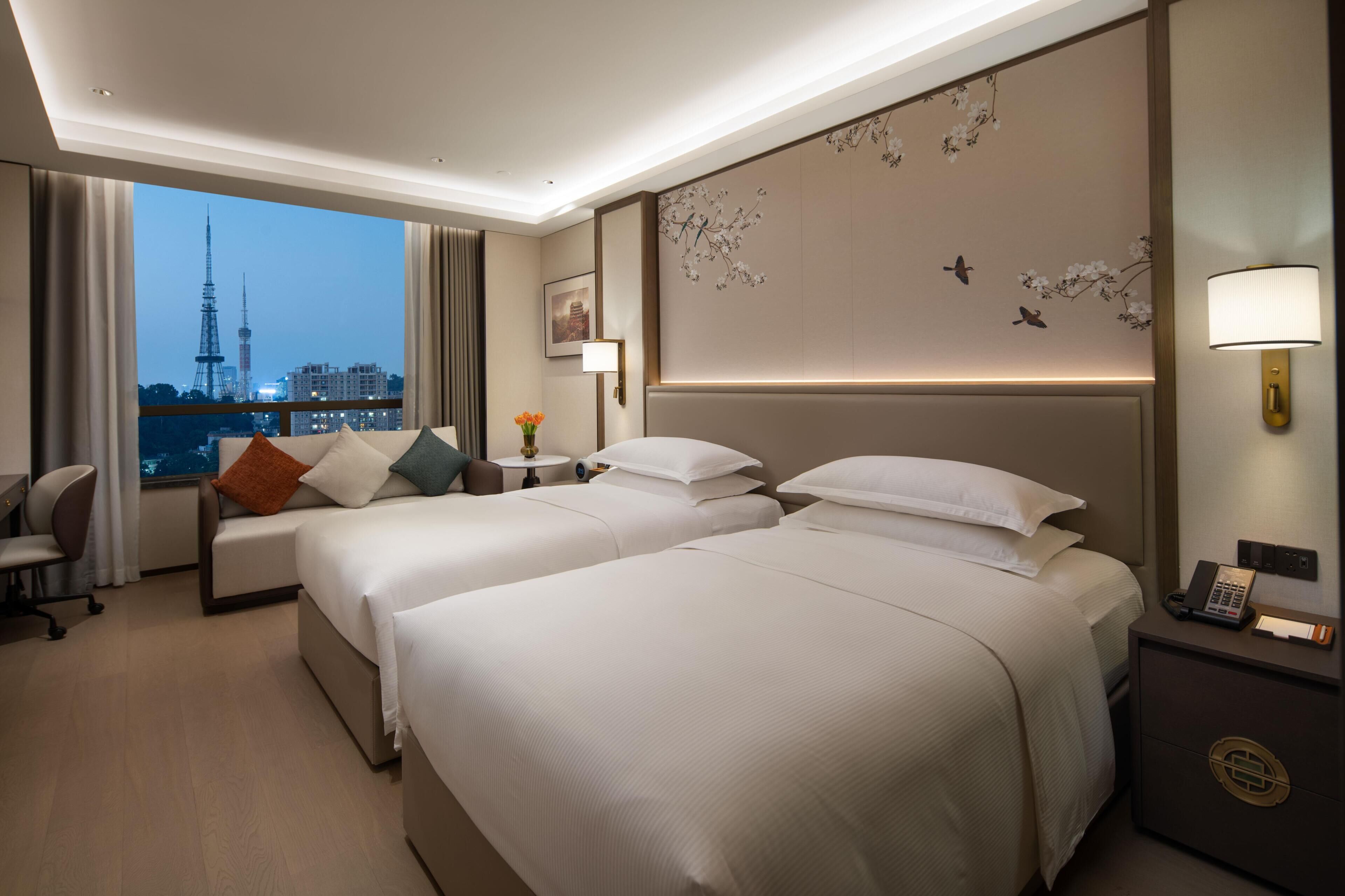A hotel room with two beds, a desk, a chair, a nightstand with a phone, and a view of a cityscape.