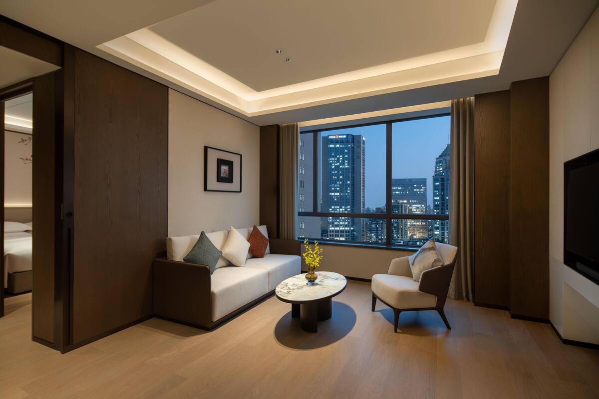 A modern hotel room with a sofa, armchair, and a coffee table. A large window offers a city view.
