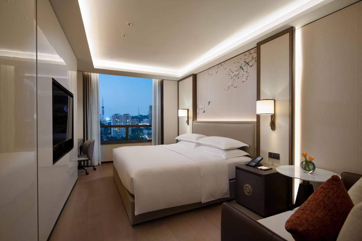 A modern hotel room with a large bed, bedside tables, a TV, and a city view.