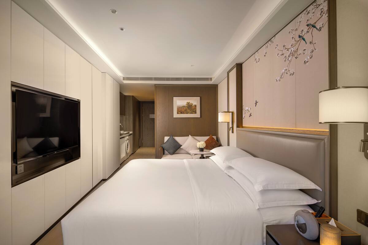 A hotel room with a large bed, a flat-screen TV, a bedside table with a lamp, and a wall-mounted TV.