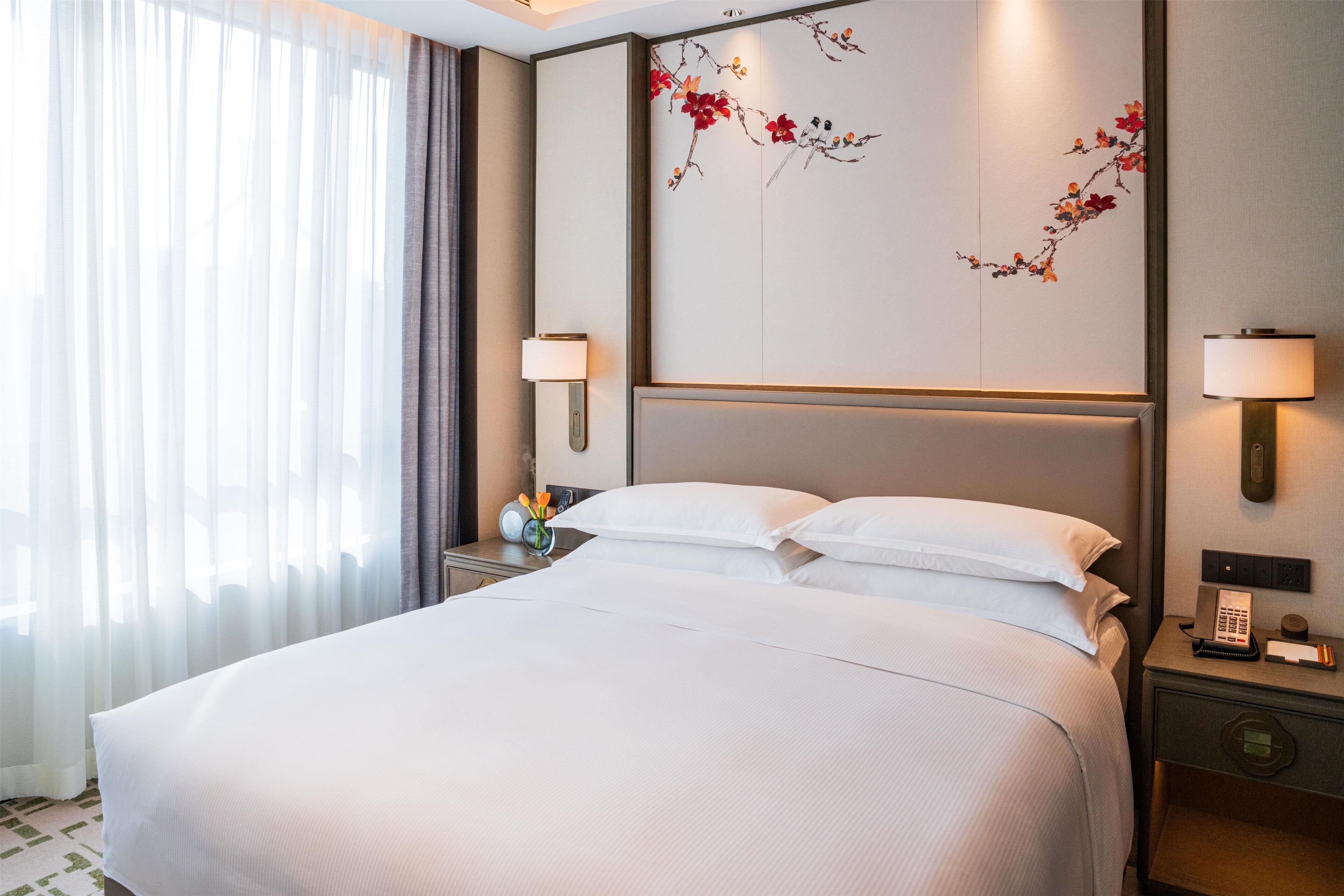 Habitación de hotel con una cama grande, dos lámparas de noche, una mesita de noche con un teléfono y una lámpara, y una pared con un cuadro floral.