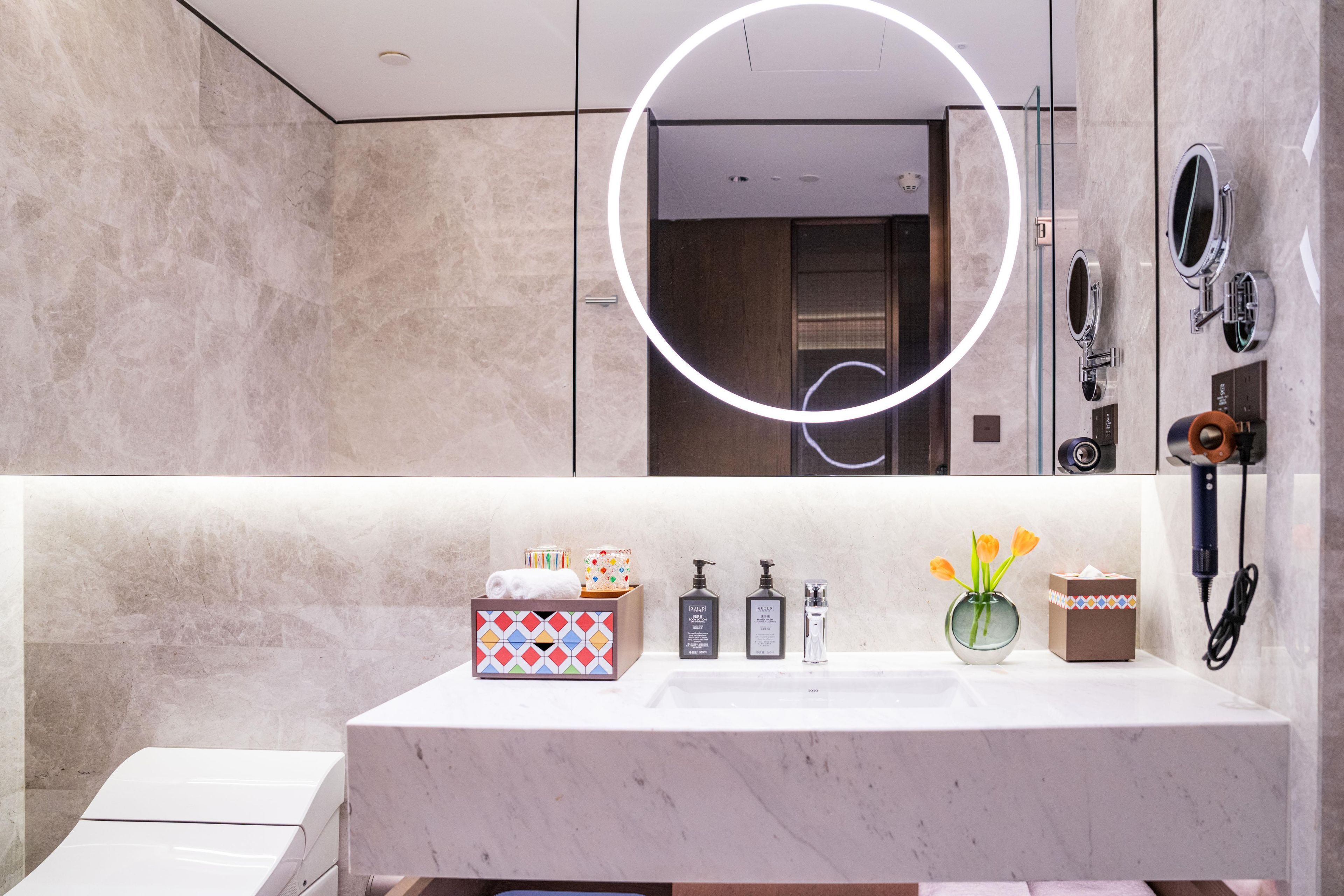 A modern bathroom with a marble sink, a mirror with integrated lighting, a handheld showerhead, and a towel dispenser.