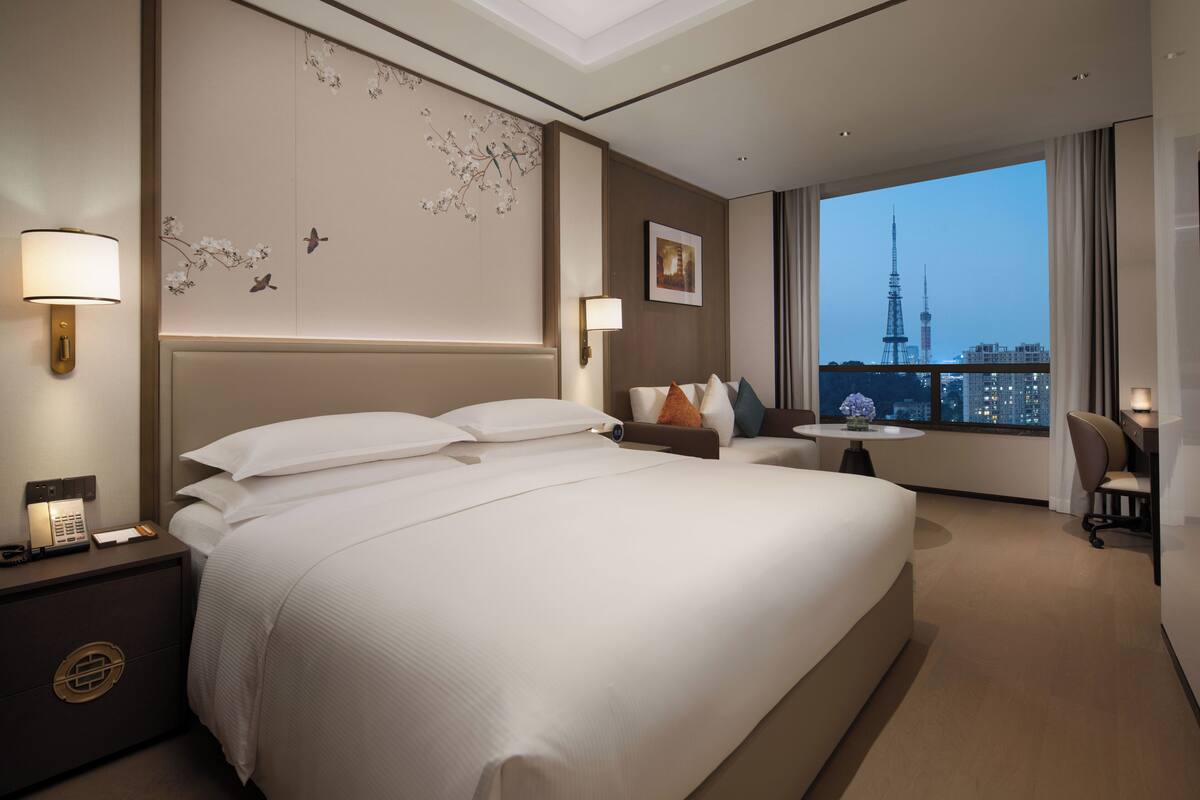 Habitación de hotel con una cama grande, lámparas de noche, una mesita con una silla y vistas a una torre.