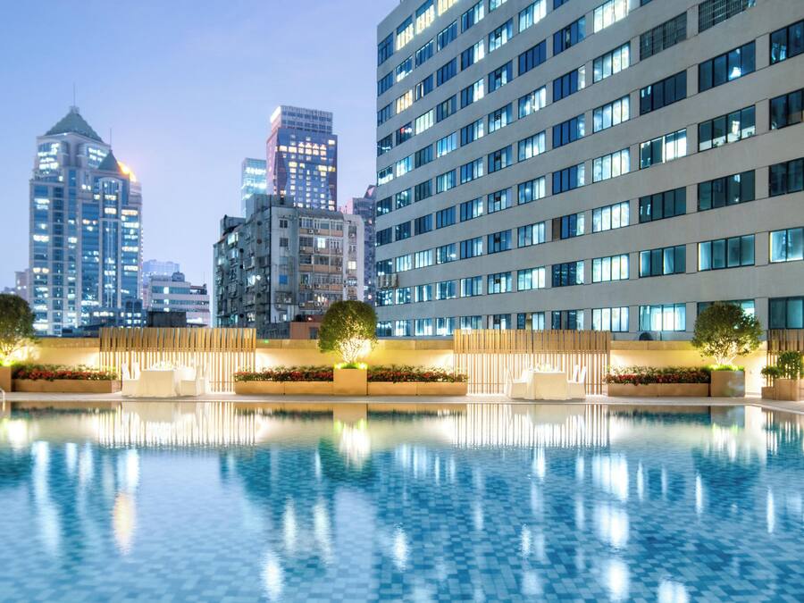 Ein modernes Hotel mit großem Pool, hoch aufragenden Gebäuden im Hintergrund und einem gut beleuchteten Außenbereich.