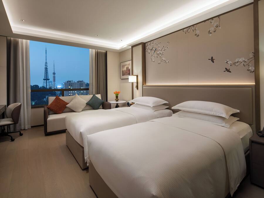 A hotel room with two beds, a desk, a chair, a nightstand, and a view of a cityscape.