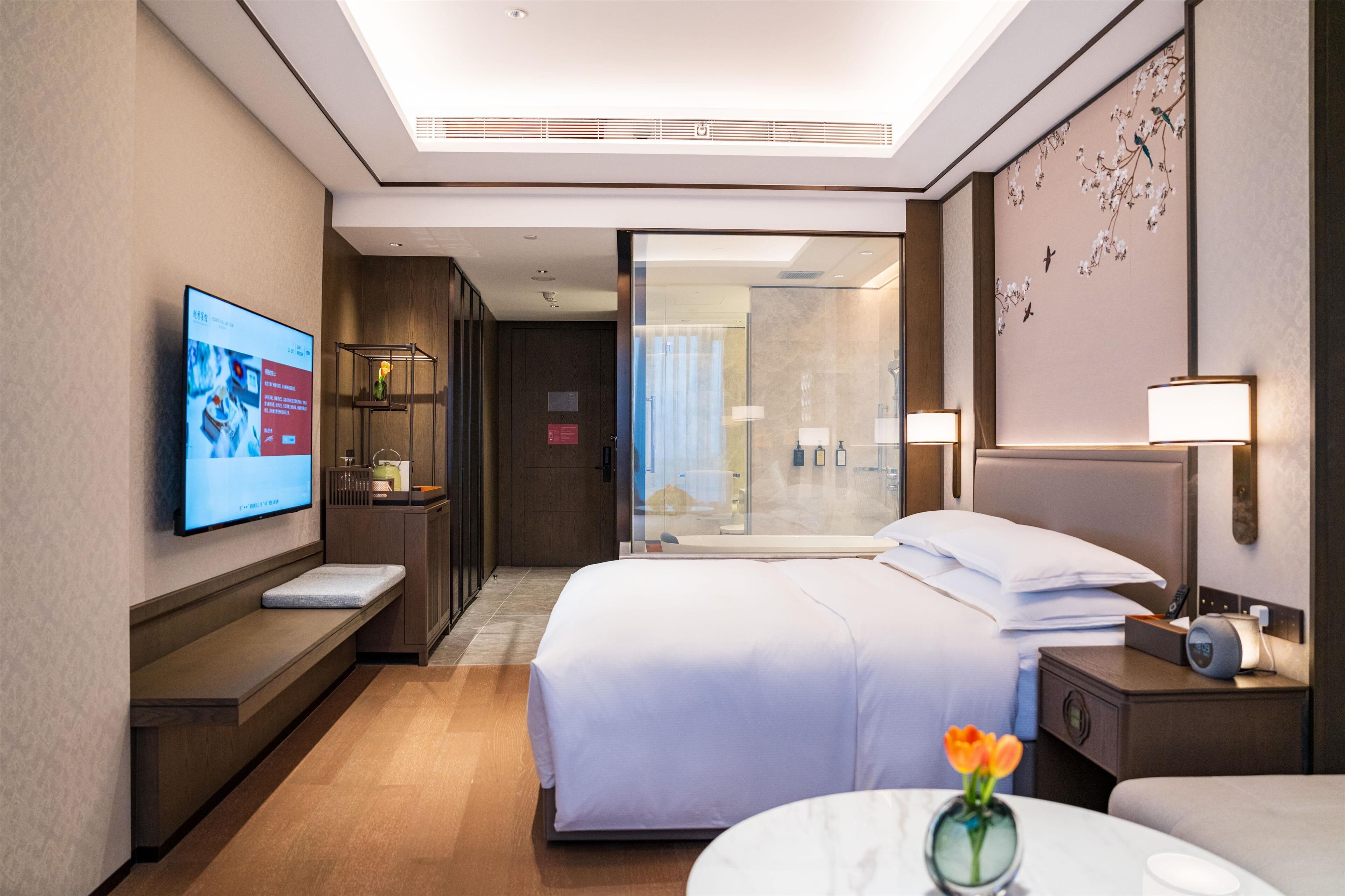 A modern hotel room with a large bed, bedside tables, a TV, and a bathroom with a glass shower.